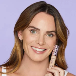 Smiling woman with green eyes and brown hair holds a Tarte Ultra Creamy concealer tube near her cheek against a lavender background.