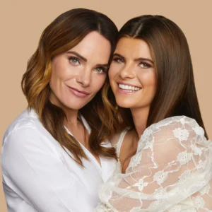 Two women posing closely together and smiling at the camera; one wears a white blouse and the other a lace wedding dress against a beige background.